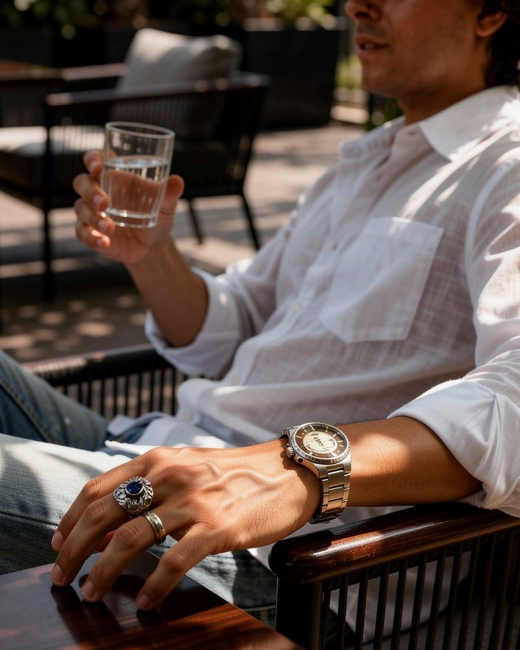 Person sitting outdoors holding a glass of water, wearing a watch and ring.